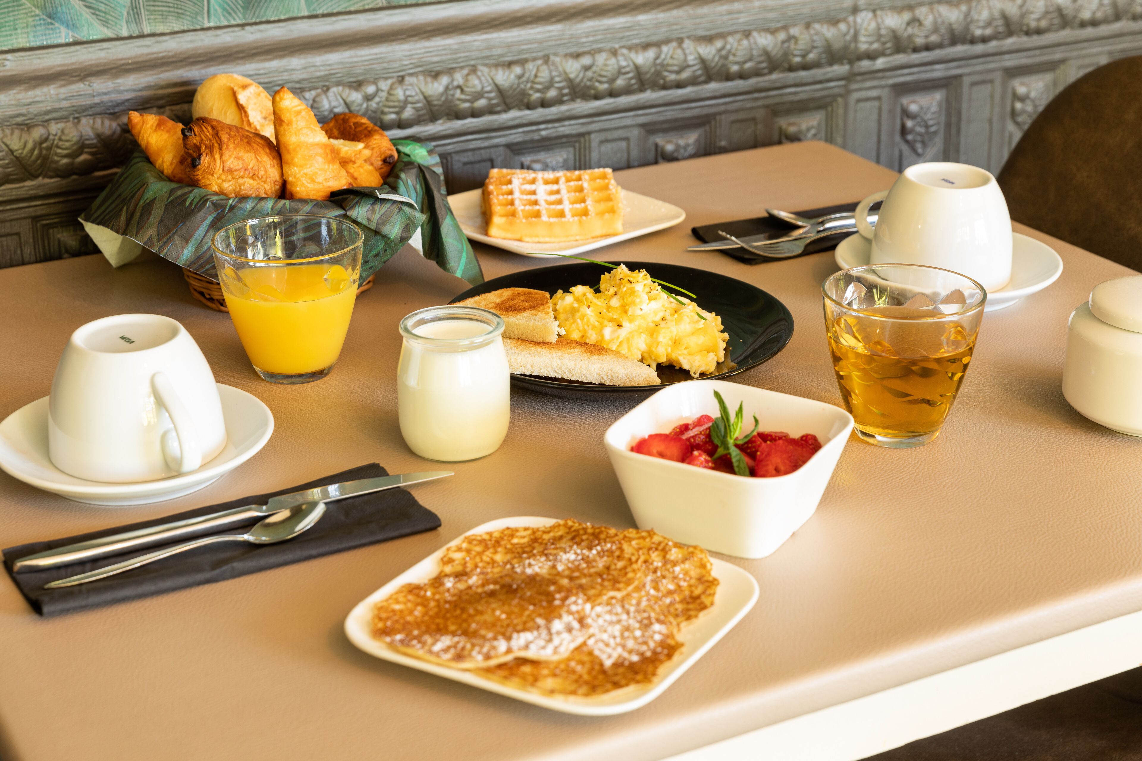 A breakfast table with croissants, waffles, scrambled eggs, toast, pancakes, a bowl of strawberries, a glass of orange juice, a glass of tea, and a cup of coffee.