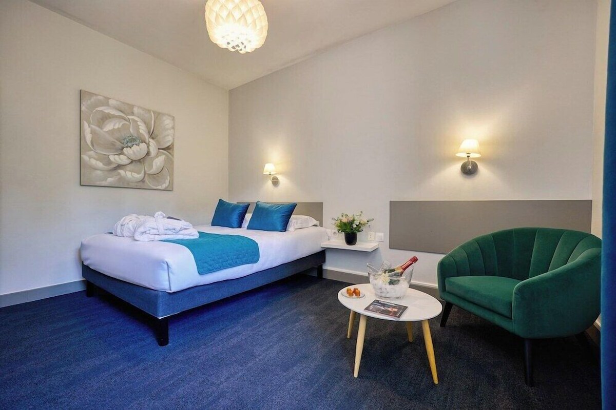 Habitación de hotel con cama, mesita de noche, lámpara, sillón verde y un cuadro en la pared.