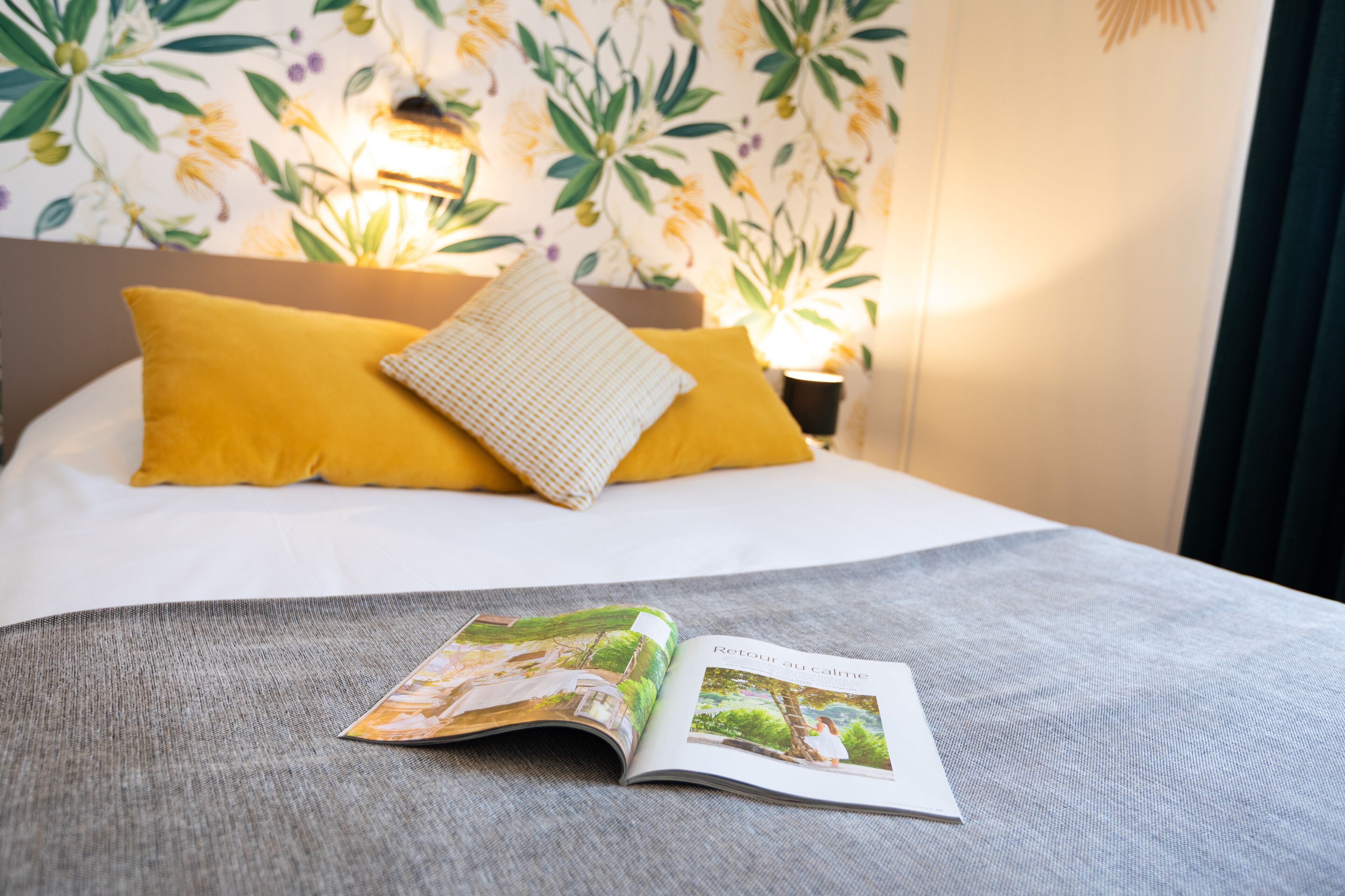 Una cama con almohadas amarillas y blancas, una revista sobre la cama, y un papel tapiz floral de fondo.