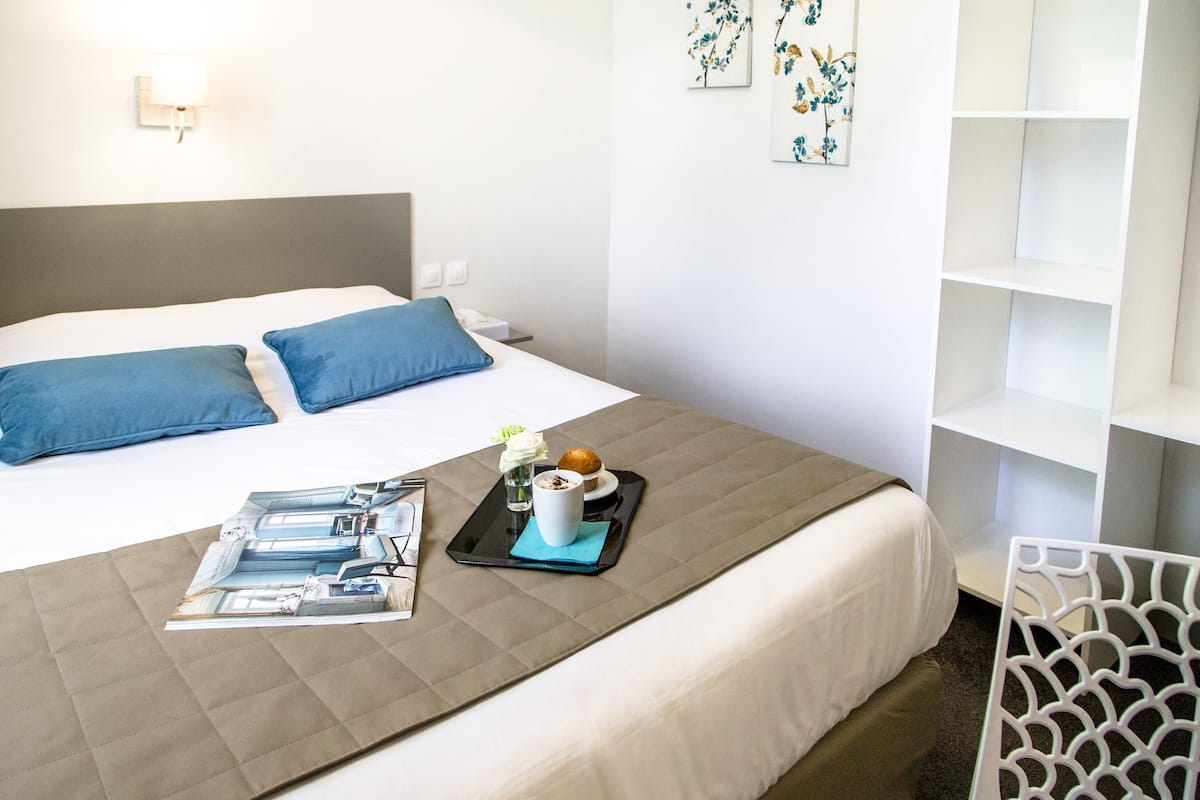 Une chambre d’hôtel avec un lit, un plateau contenant un muffin et des fleurs, un magazine et une chaise blanche.