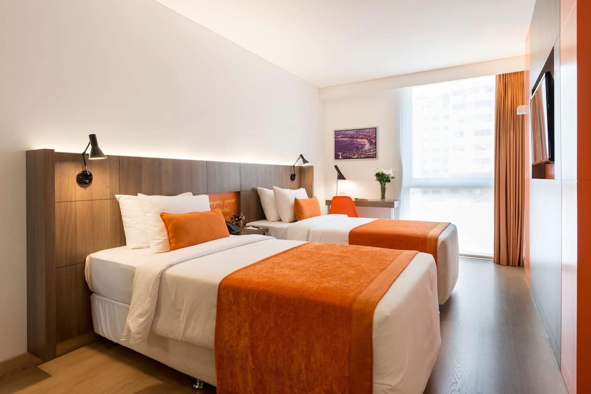 A hotel room with two beds, a wooden headboard, a nightstand, a desk, a chair, and a large window with curtains.