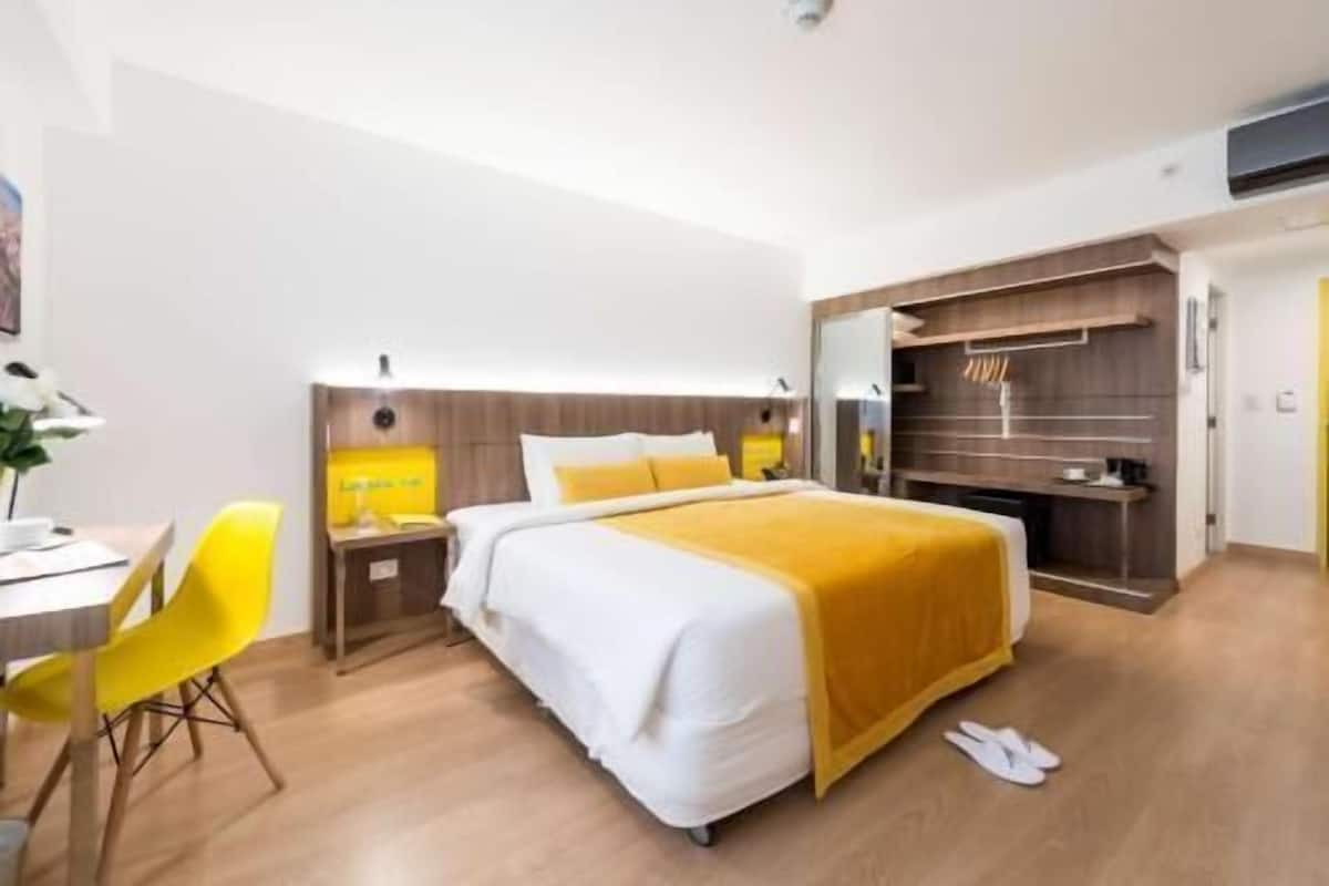 A modern hotel room with a large bed, a desk, a chair, and a small kitchenette area.