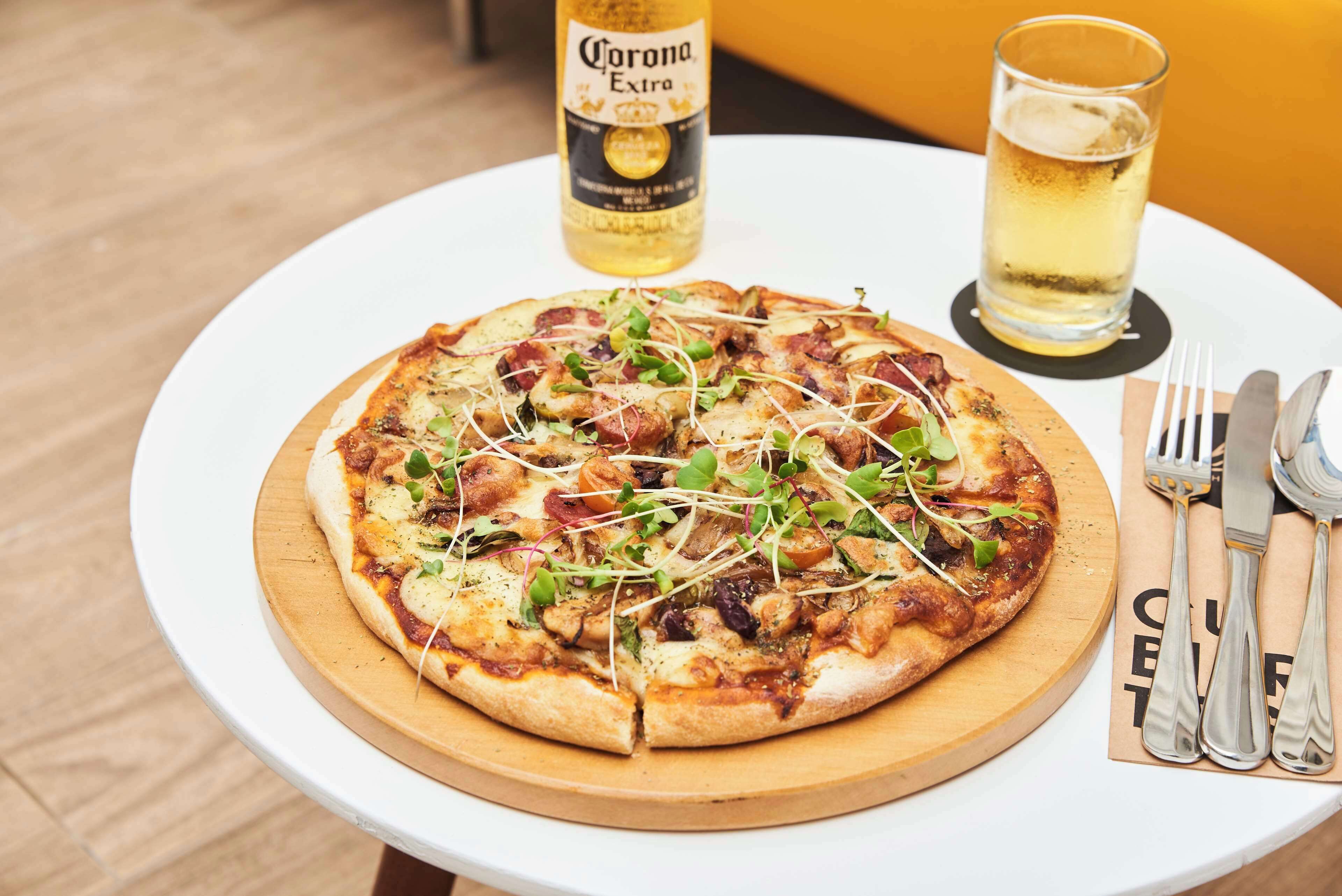 Une pizza servie sur une planche en bois, accompagnée d’une bouteille de bière Corona Extra et d’un verre de bière, le tout posé sur une table blanche.