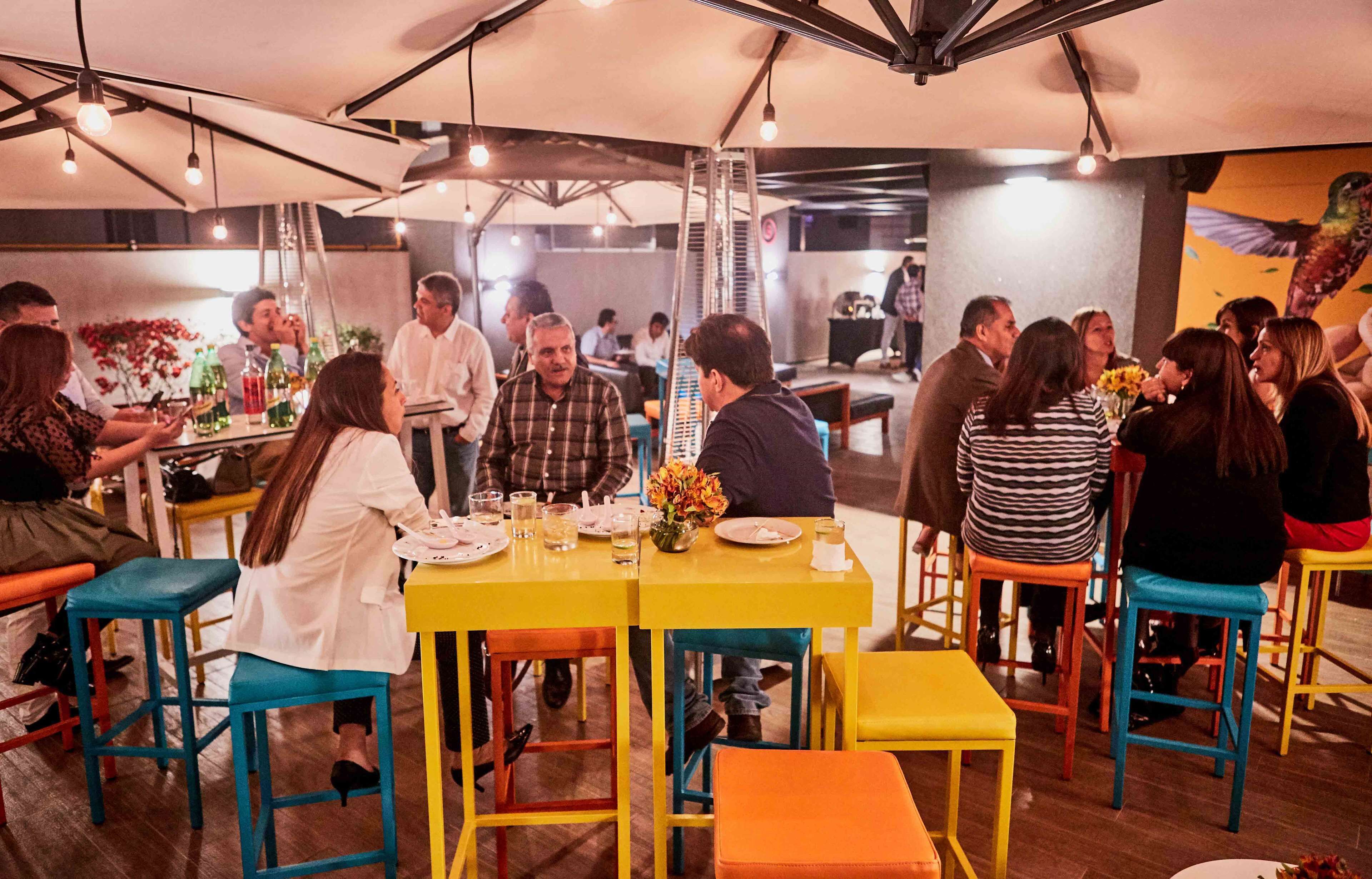Reunión social en un restaurante con mesas amarillas y taburetes coloridos, rodeado de comensales conversando.