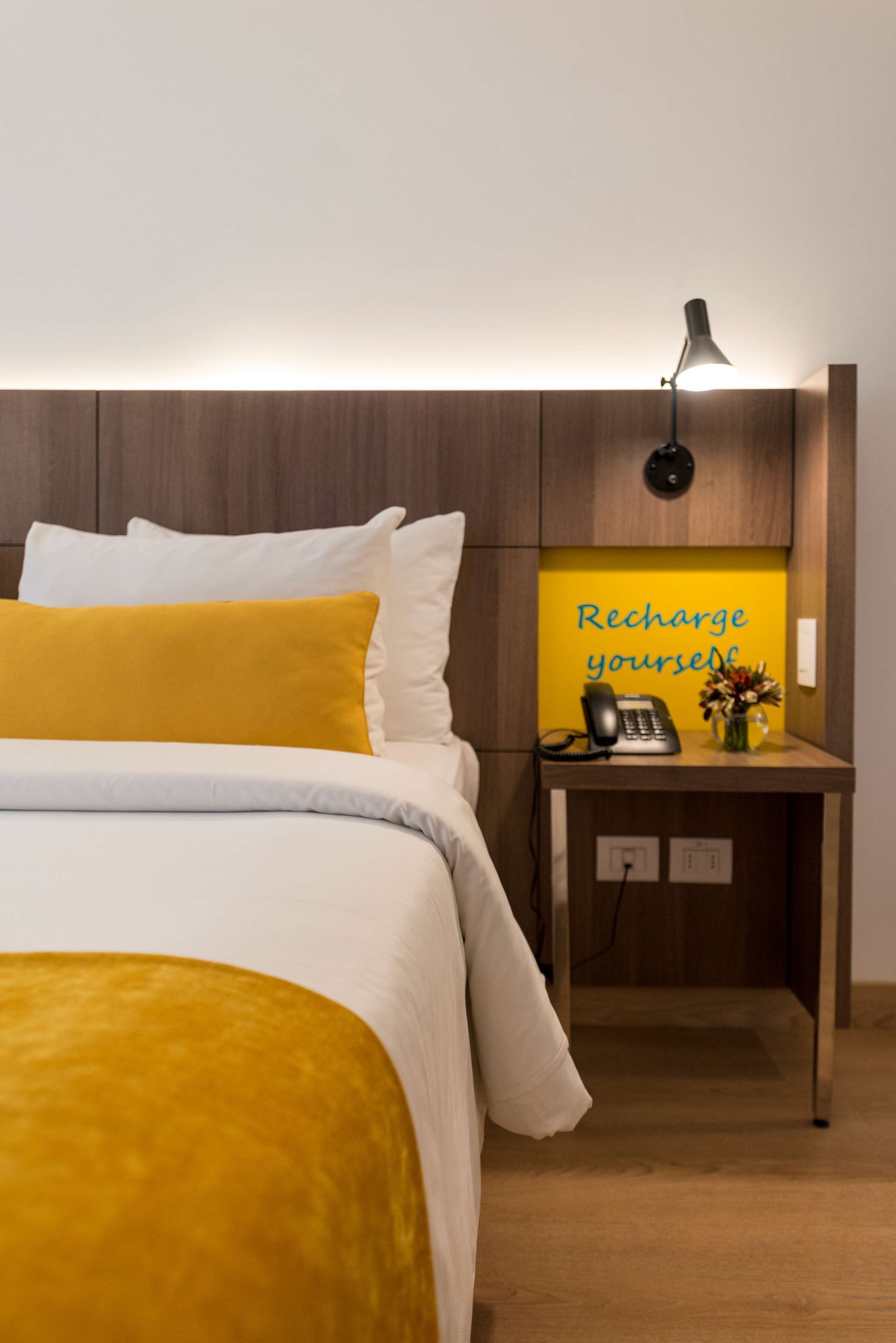 Een hotelkamer met een bed, een nachtkastje met een lamp, een telefoon en een bordje met de tekst 
