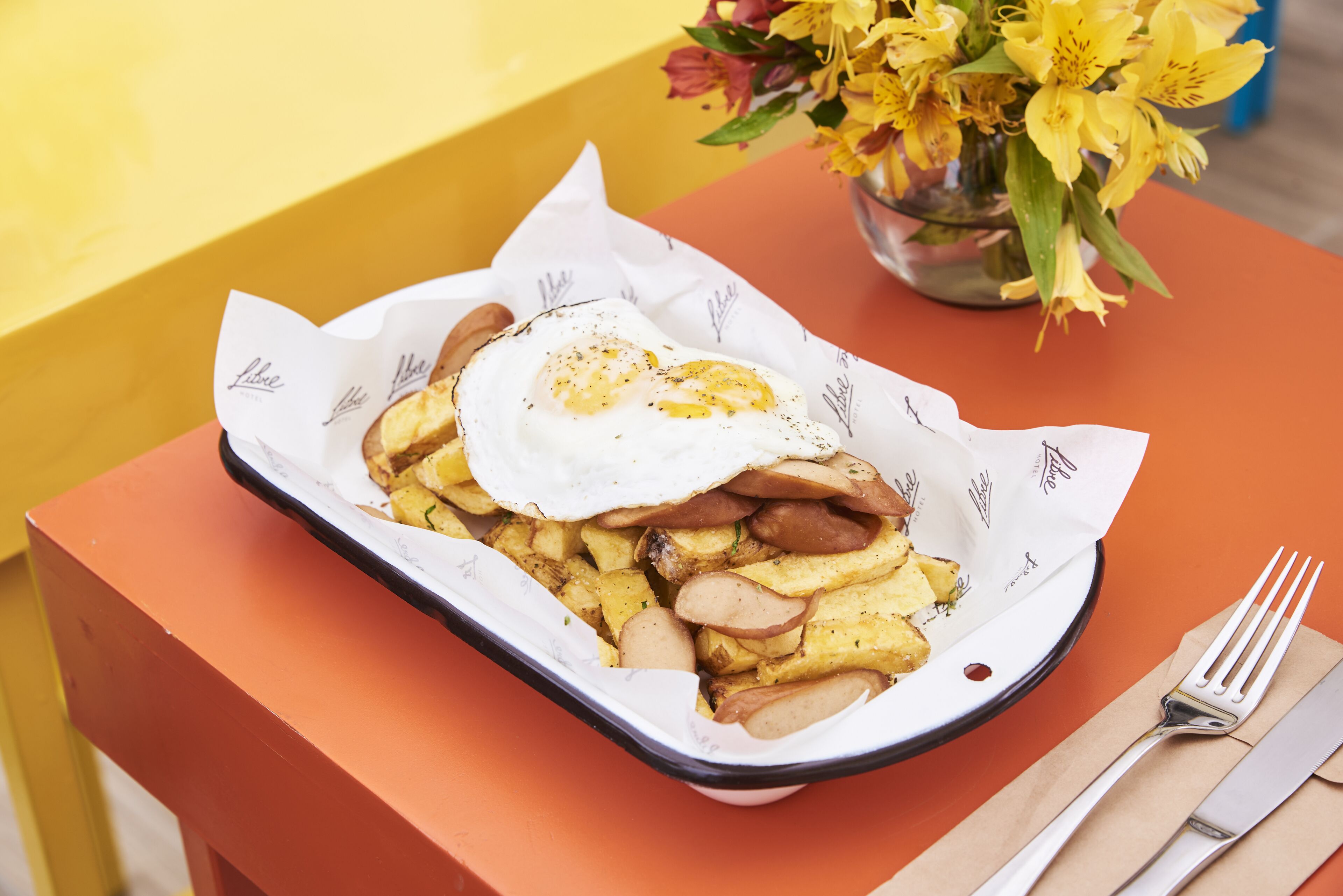 A tray of food with a fried egg and potatoes on a table with a vase of flowers.