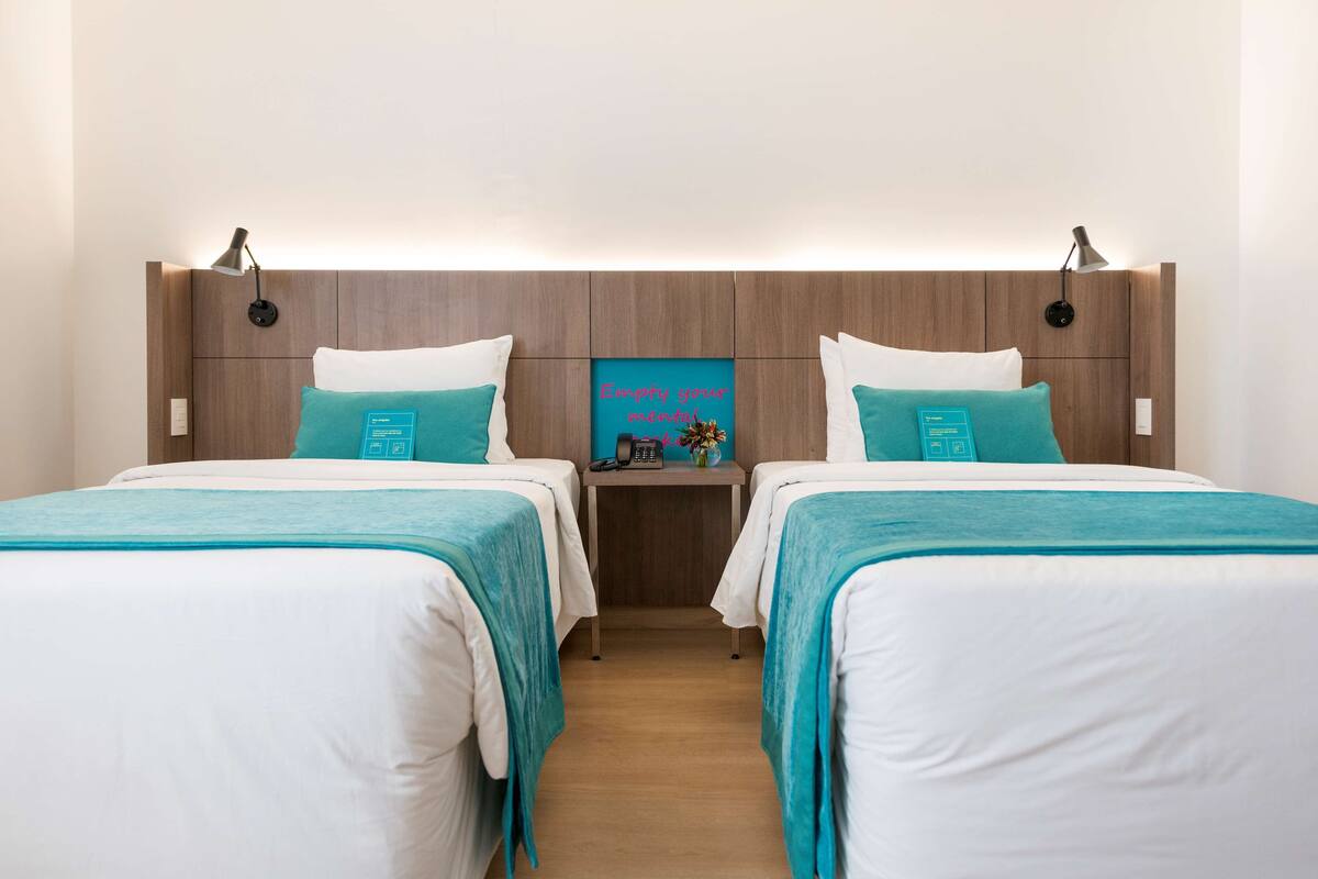 A hotel room with two beds, each with white and teal bedding, a wooden headboard, and a small table with a lamp and a sign that reads "Empty your mind, fill your heart."