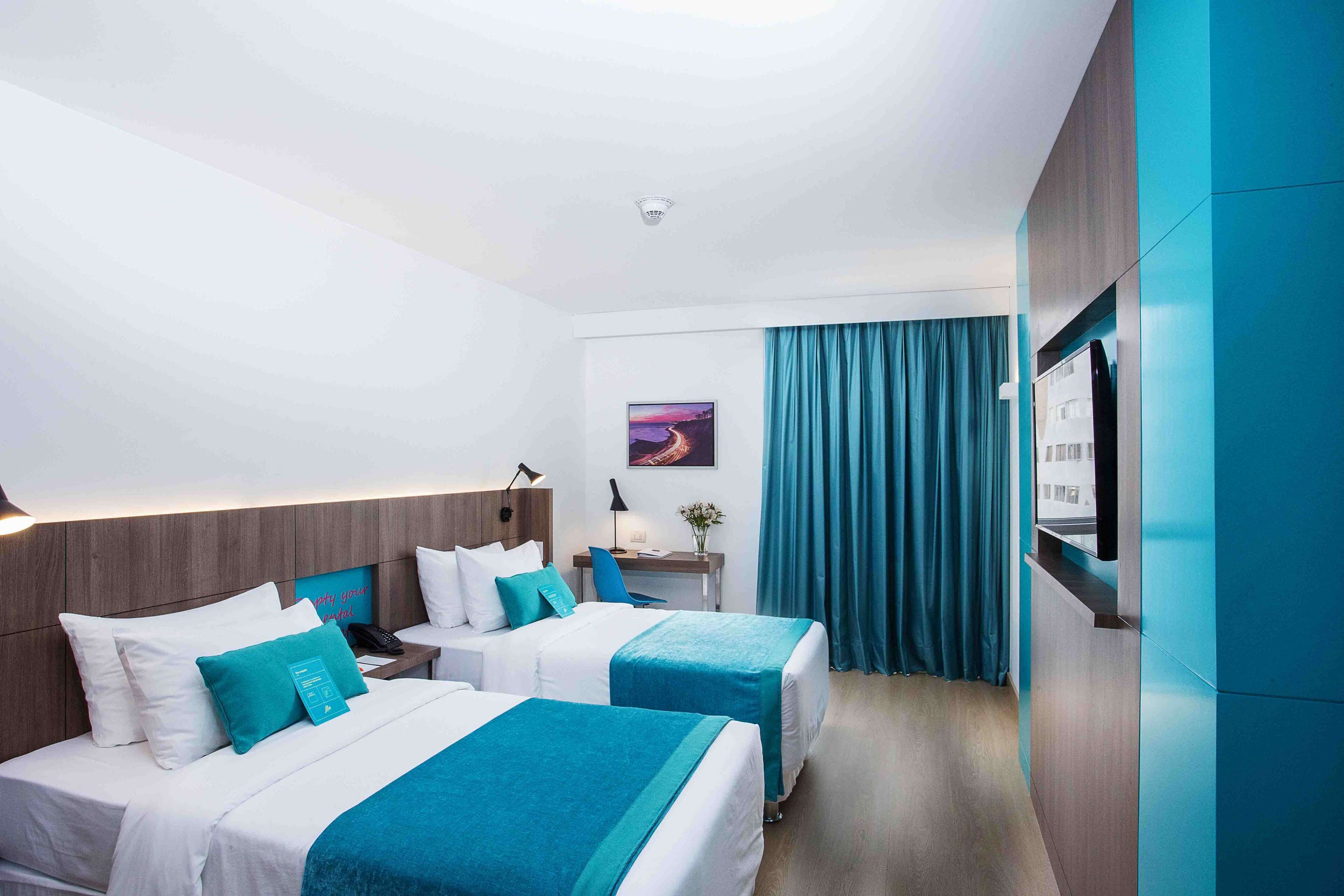Ein Hotelzimmer mit zwei Betten, einem Schreibtisch, einem Stuhl, einem Fernseher und einem Fenster mit Vorhängen.