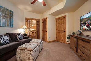 A living room with a sofa, ottomans, a lamp, a ceiling fan, and a television on a cabinet. - 4201 Aspen Lodge, Trappeur's Crossing Apartment 3 (Steamboat Springs)