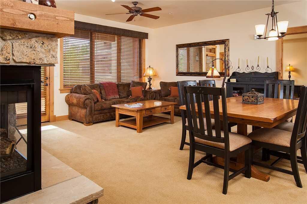A cozy living room with a fireplace, a wooden dining table with chairs, a sofa, and a ceiling fan.