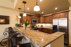 A kitchen with granite countertops, wooden cabinets, a stainless steel refrigerator, and pendant lighting.