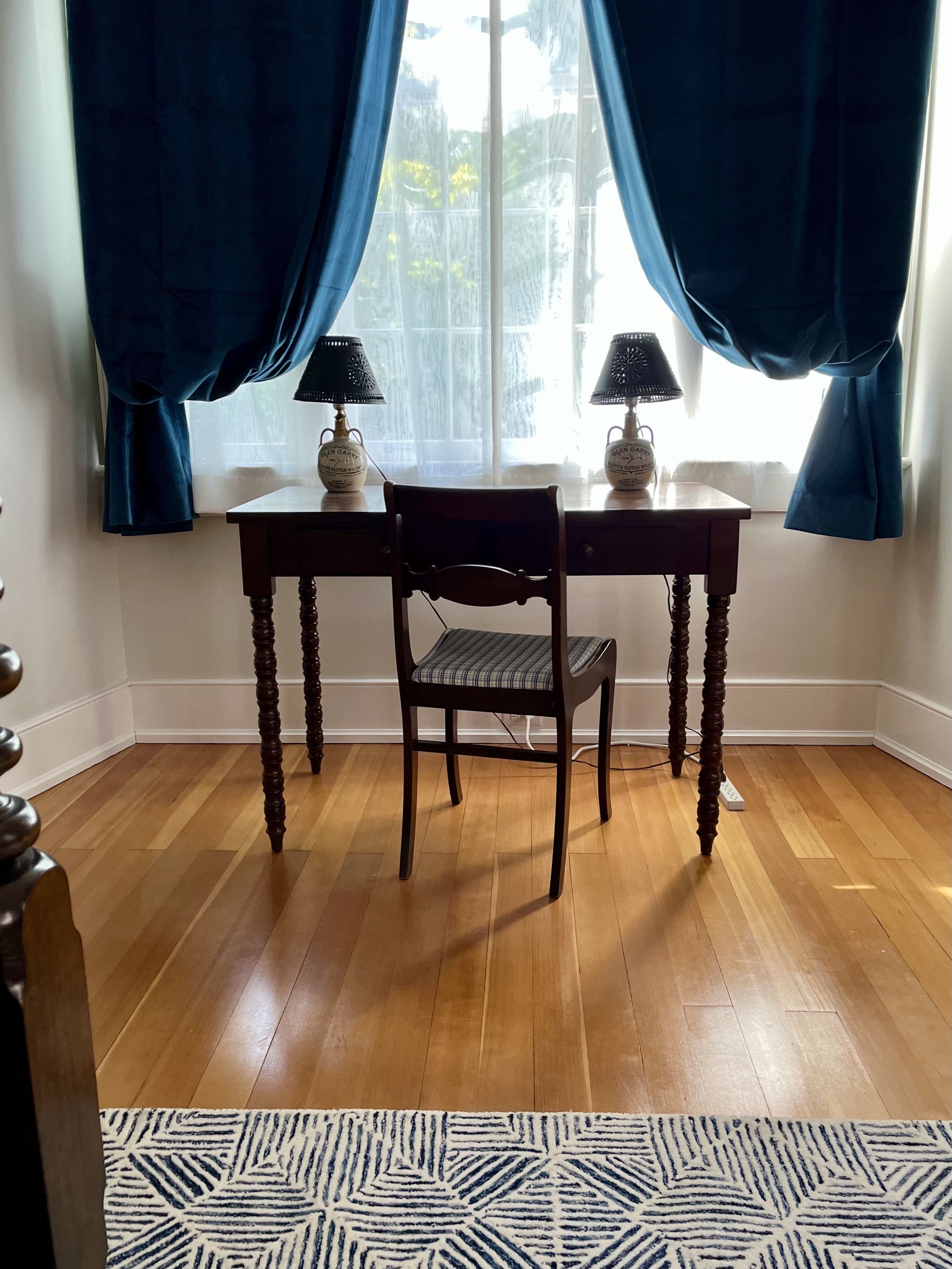 A room with a wooden floor, a table with two lamps, a chair, and a window with curtains.