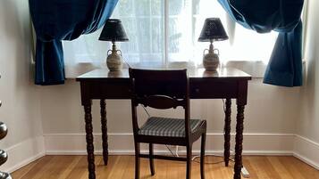 A room with a wooden floor, a table with two lamps, a chair, and a window with curtains.