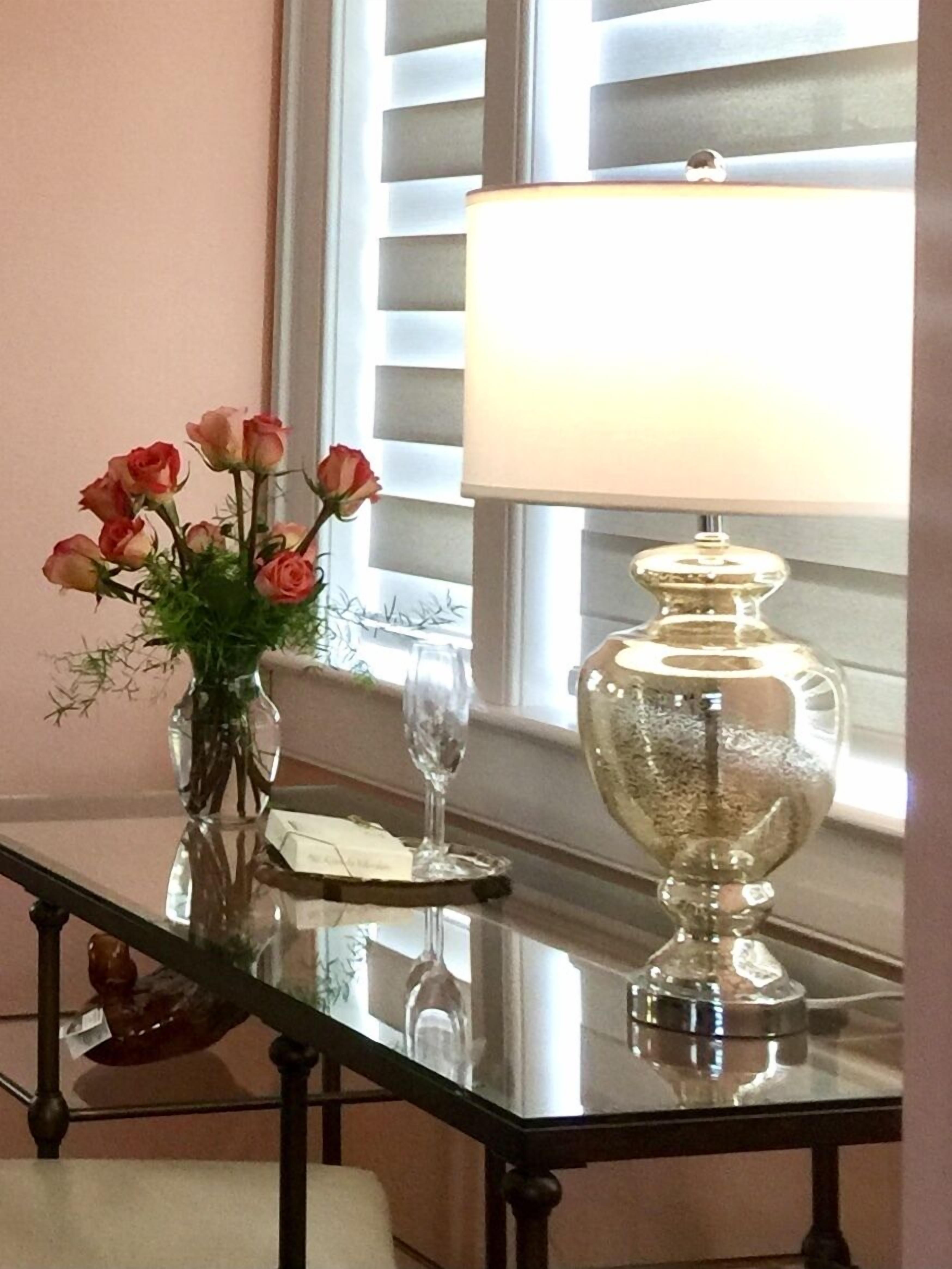 Une table en verre sur laquelle se trouve un vase rempli de roses roses, un verre à vin transparent et un bol décoratif en argent.