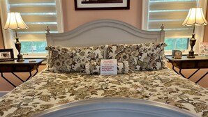 A neatly made bed with floral bedding, two bedside tables with lamps, a framed picture on the left table, and a welcome sign on the bed. - Charred Oaks Inn (Versailles)