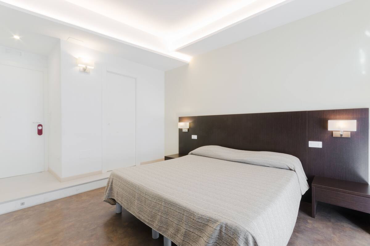 A modern hotel room with a large bed, bedside tables, a wardrobe, and recessed ceiling lighting.
