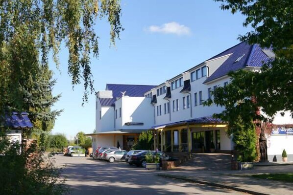 A hotel with a white facade, multiple windows, and a blue roof. There are parked cars in front and a person standing near the entrance. - Erbenholz Hotel (Laatzen)