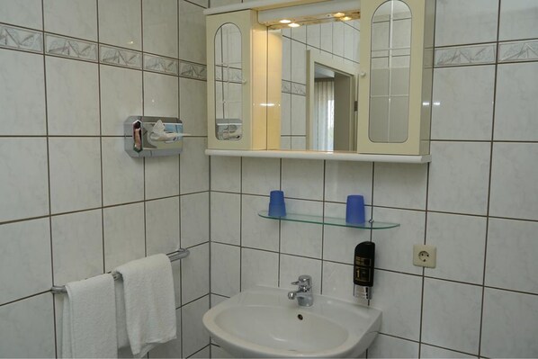 Un baño con un lavamanos blanco, un espejo y un estante de vidrio con artículos de aseo.