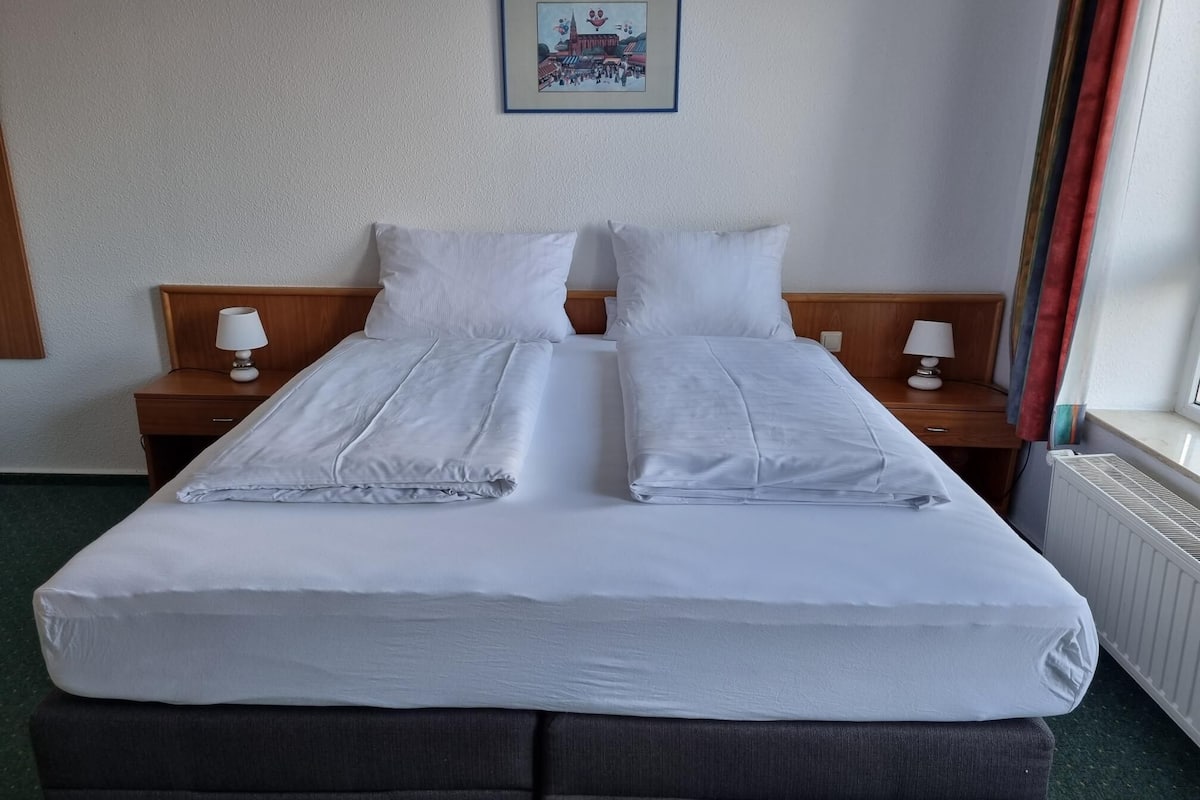 A hotel room with a double bed, two bedside tables with lamps, a framed picture on the wall, and a window with curtains.