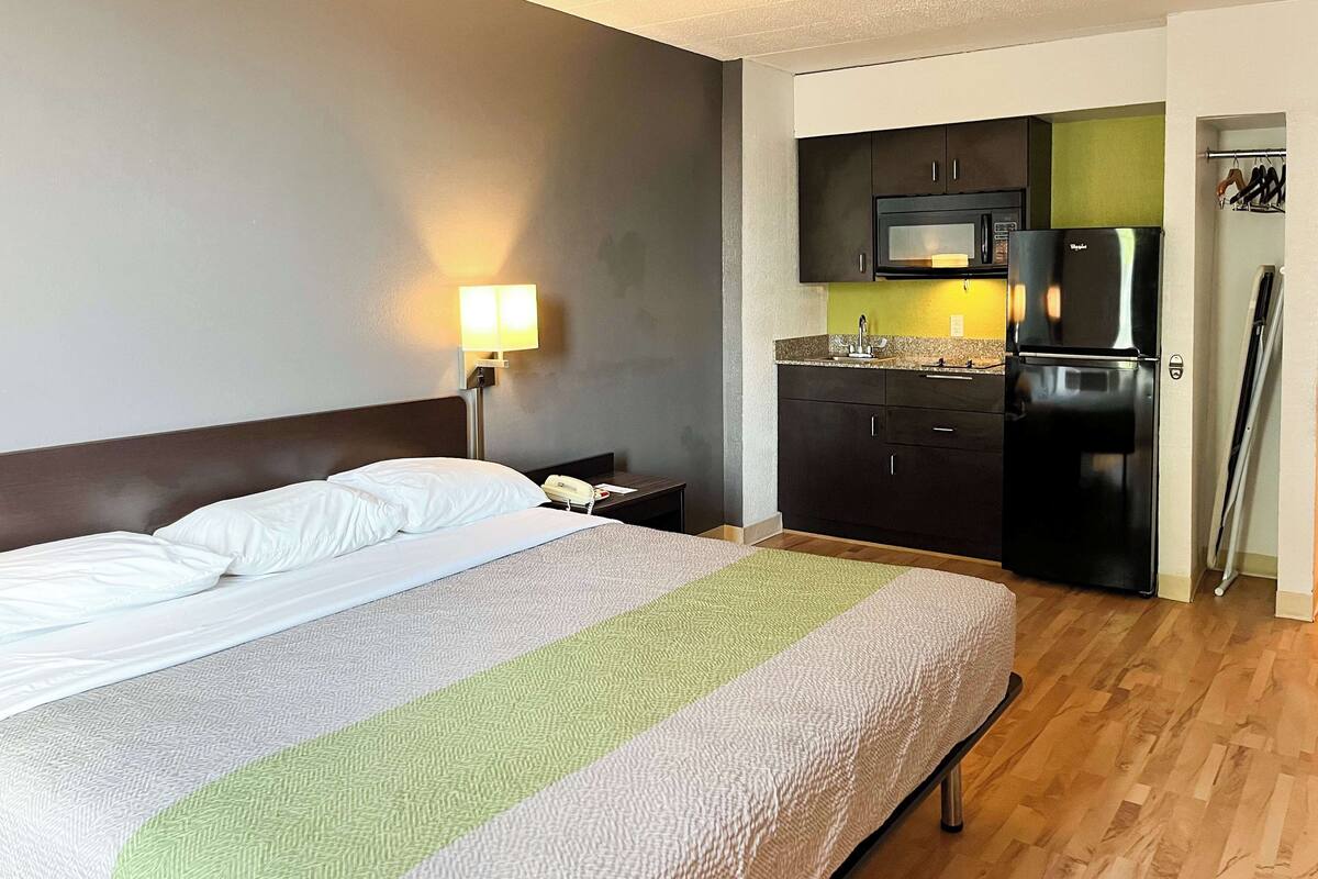 A hotel room with a bed, a nightstand, a lamp, a kitchenette with a microwave and refrigerator, and a wooden floor.