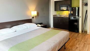 Habitación de hotel con cama, mesita de noche, lámpara, kitchenette con microondas y refrigerador, y piso de madera.
