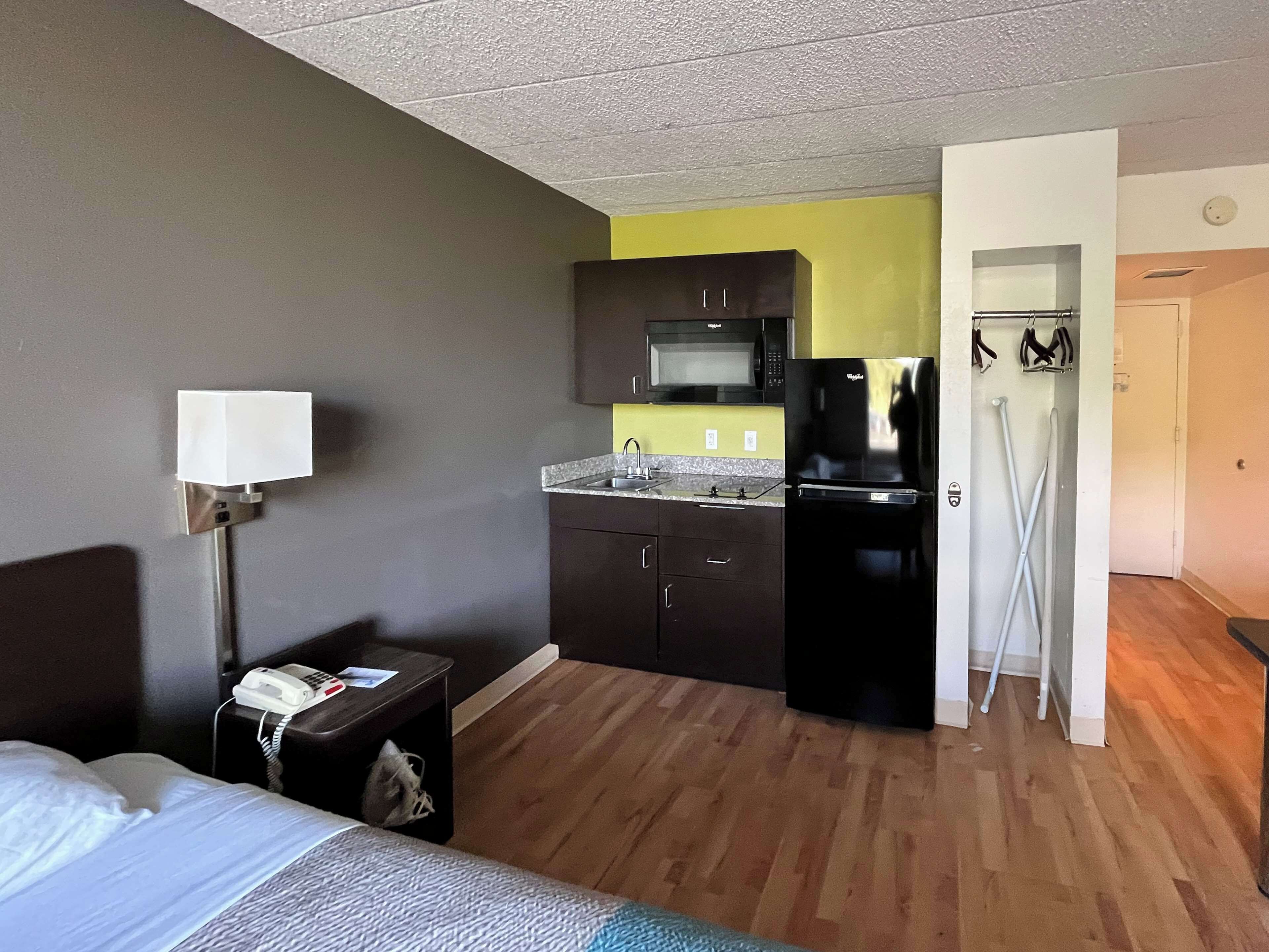 A hotel room with a bed, a bedside table with a lamp, a kitchenette with a microwave, a refrigerator, and a door leading to a bathroom.