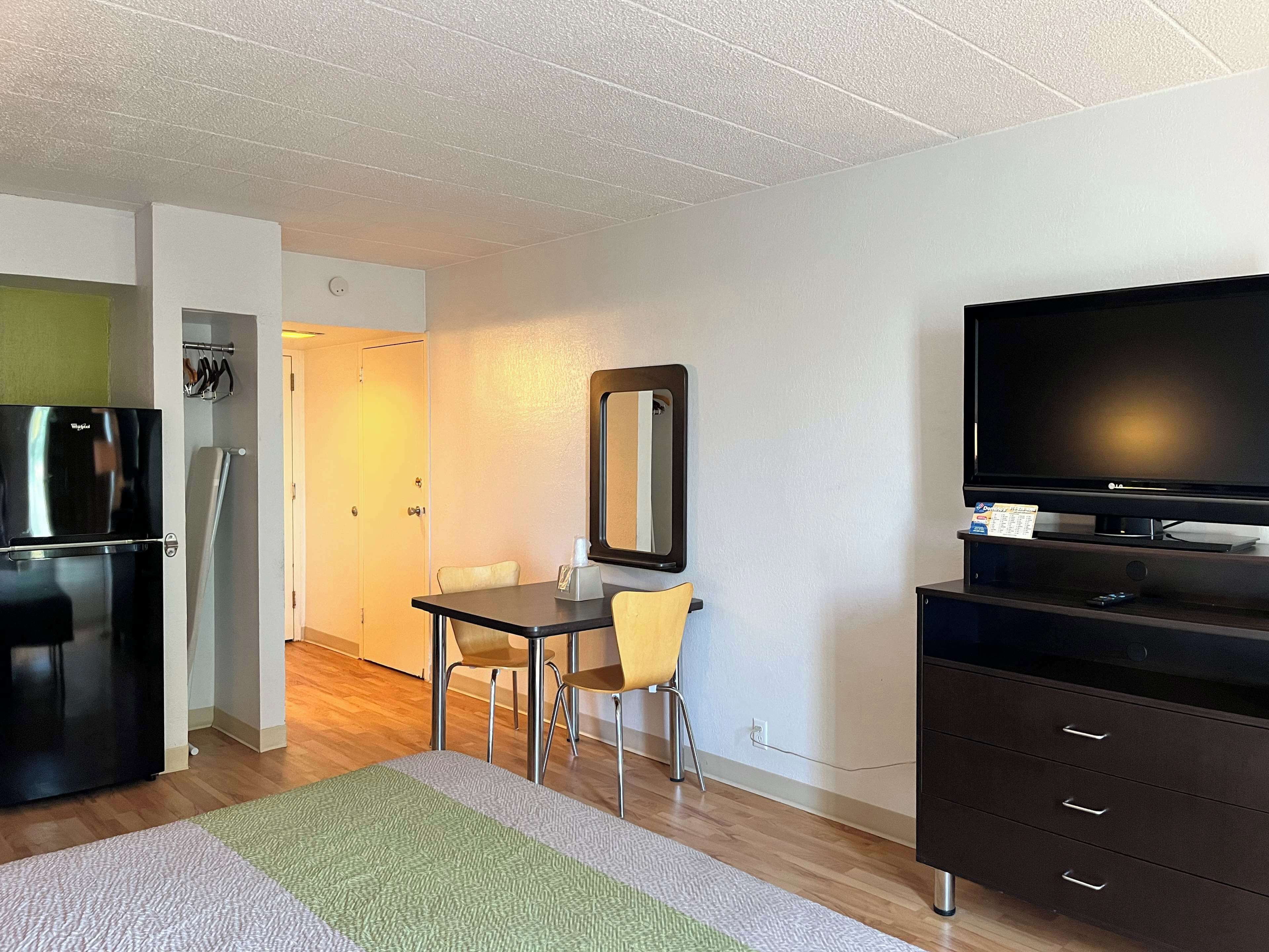 A compact hotel room with a kitchenette, a dining area with a table and chairs, a large mirror, a flat-screen TV, and a dark-colored dresser.