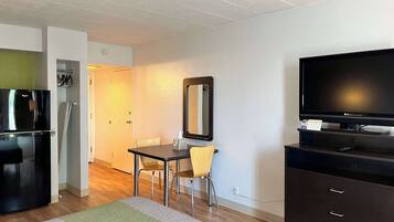 A compact hotel room with a kitchenette, a dining area with a table and chairs, a large mirror, a flat-screen TV, and a dark-colored dresser.