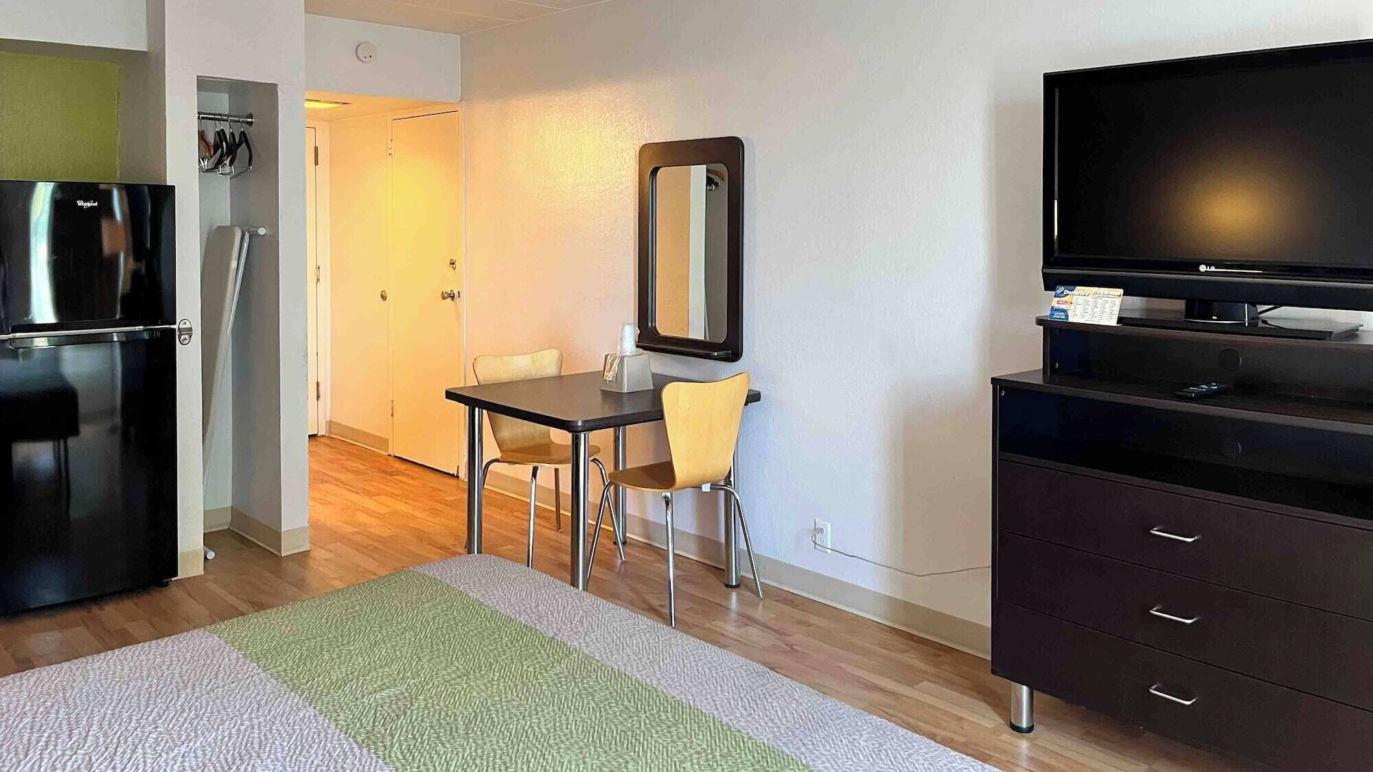 A compact hotel room with a kitchenette, a dining area with a table and two chairs, a large mirror, a flat-screen TV, and a dark-colored dresser.