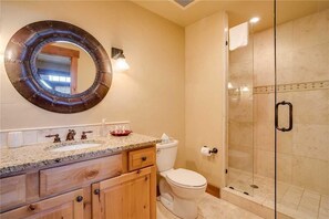 A bathroom with a round mirror, a sink with a granite countertop, wooden cabinets, a toilet, and a glass-enclosed shower. - 5114 Emerald Lodge, Trappeur's Crossing Apartment 1 (Steamboat Springs)