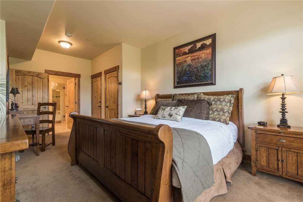 A bedroom with a wooden bed, a desk, and a chair. There is a painting on the wall and a lamp on the bedside table.
