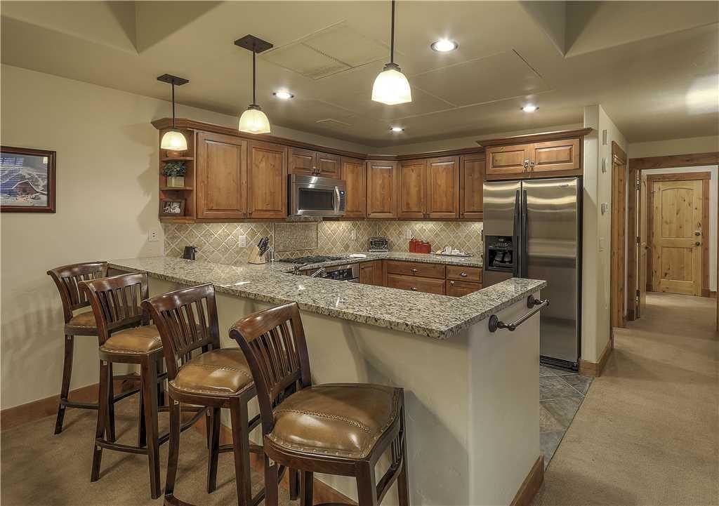 A kitchen with wooden cabinets, stainless steel appliances, and a granite countertop. There are four bar stools and pendant lights.