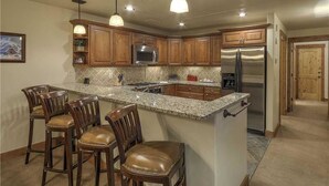 Cocina con armarios de madera, electrodomésticos de acero inoxidable y encimera de granito. Hay cuatro taburetes de bar y luces colgantes.