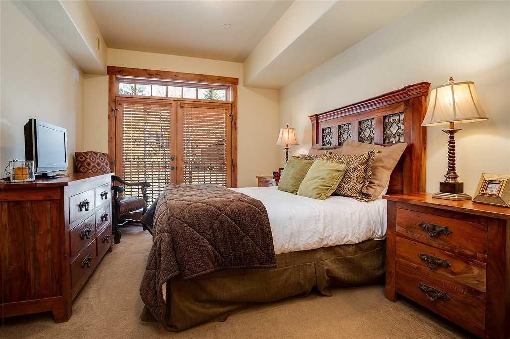 A bedroom with a bed, wooden headboard, bedside lamps, a dresser, a television, and a window with blinds.
