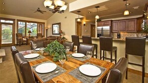 Uma sala de jantar com uma mesa de madeira posta para seis pessoas, rodeada por cadeiras de couro. Há um ventilador de teto e iluminação pendente.