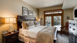 A bedroom with a large bed, a nightstand with a lamp, a dresser, and a window with a view of trees. - 5306 Emerald Lodge, Trappeur's Crossing Apartment 4 (Steamboat Springs)