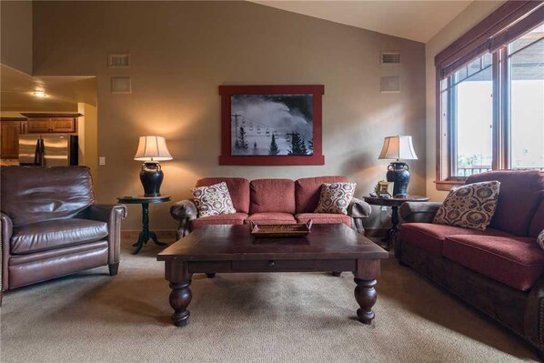 A living room with a dark wooden coffee table, two red sofas with patterned cushions, a brown leather armchair, and a framed picture on the wall.