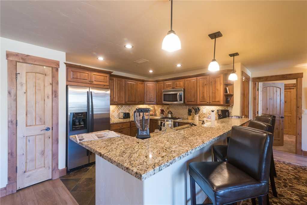 A kitchen with wooden cabinets, a granite countertop, a refrigerator, a microwave, a blender, and a bar area with chairs.