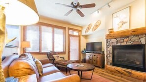 A cozy living room with a stone fireplace, a ceiling fan, and a large TV. - 2106 Timberline Lodge, Trappeur's Crossing Apartment 2 (Steamboat Springs)