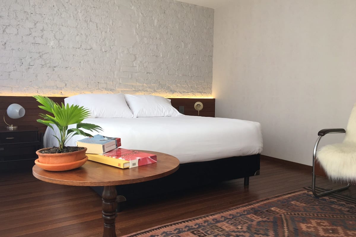 Ein modernes Schlafzimmer mit Ziegelwand, einem ordentlich bezogenen Bett, einem kleinen runden Tisch mit Pflanze und Büchern sowie einem Stuhl mit flauschigem Sitz.