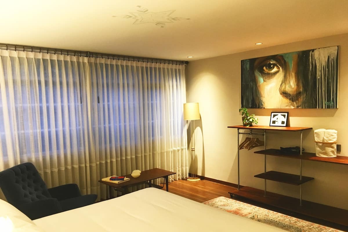 Ein Hotelzimmer mit einem Bett, einem Schreibtisch, einem Sessel, einer Lampe, einem Gemälde und einem Regal mit Pflanze und Bilderrahmen.