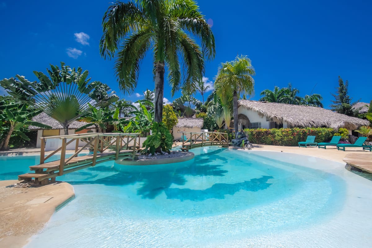 A resort pool area with clear blue water, wooden deck, palm trees, and thatched-roof huts.