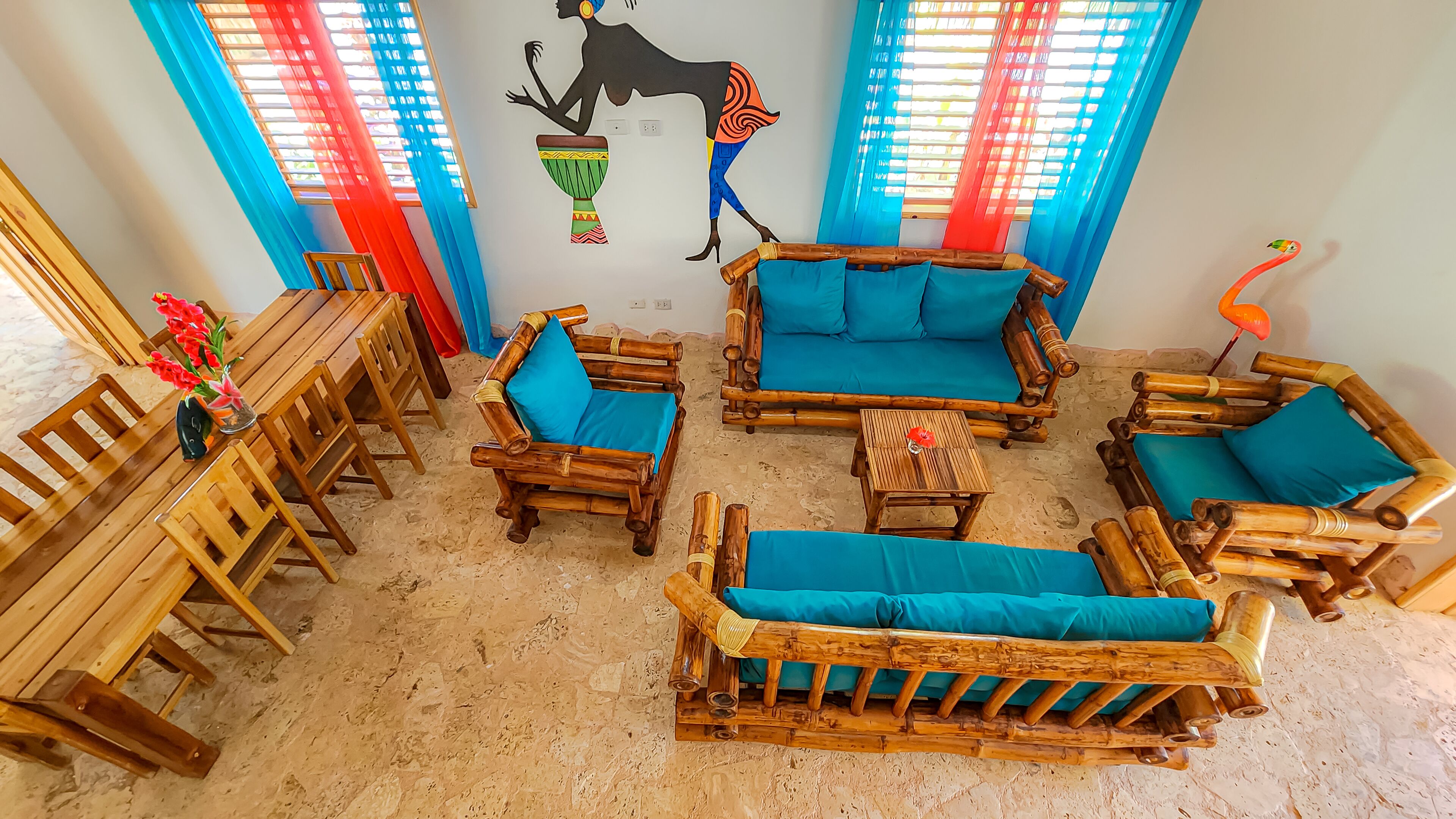 A room with a wooden dining table, chairs, and a sofa set. There is a wall mural and a window with blue curtains.