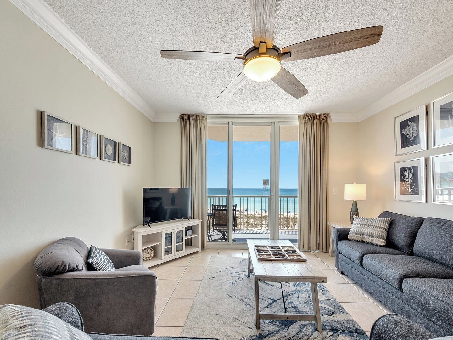Una sala de estar con ventilador de techo, televisor, sofá, mesa de centro y vista a la playa a través de una puerta de vidrio.