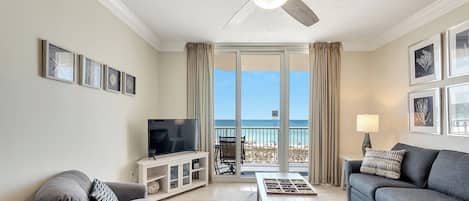 Una sala de estar con ventilador de techo, televisor, sofá, mesa de centro y vista a la playa a través de una puerta de vidrio.
