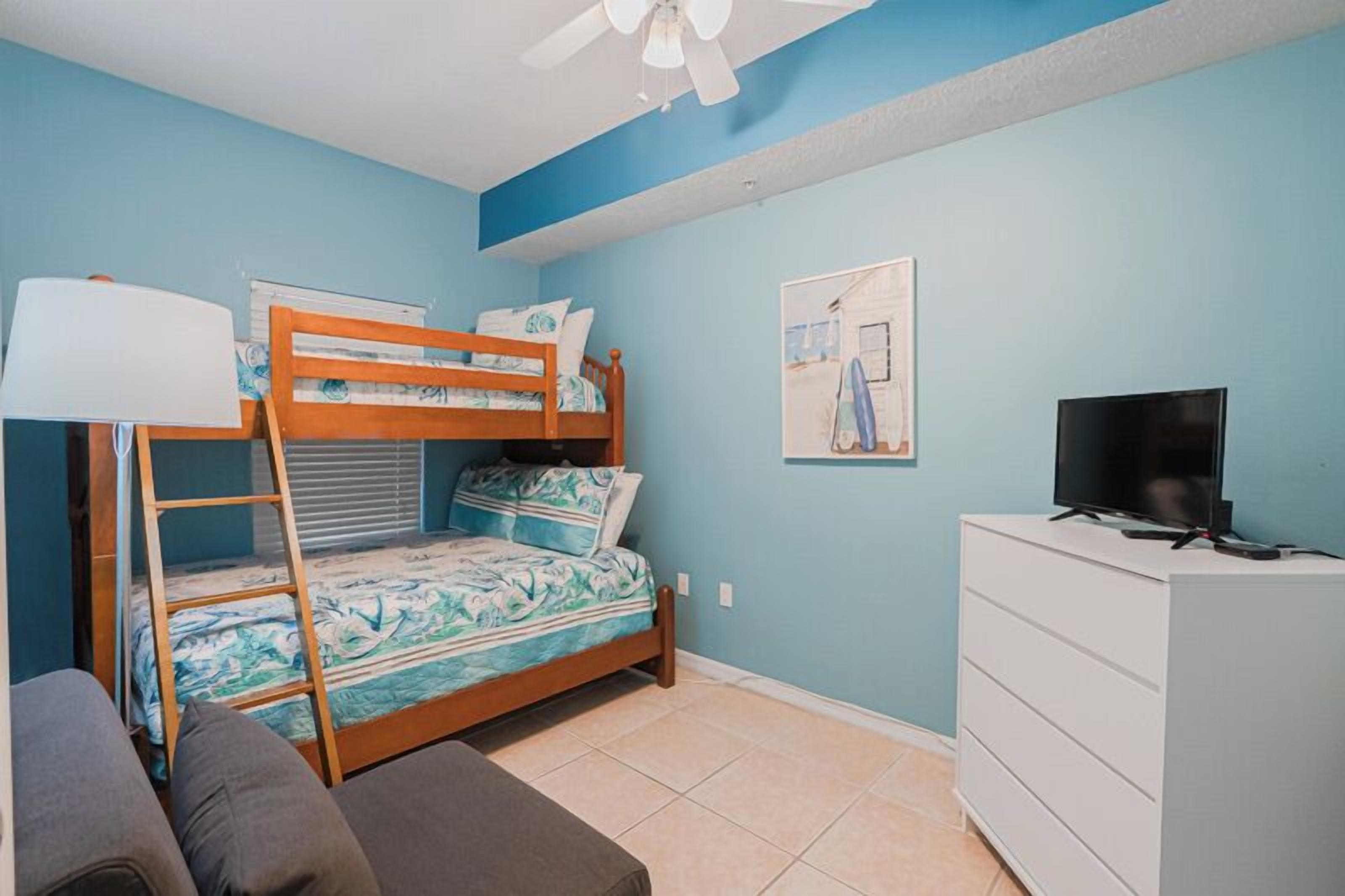 A bedroom with a bunk bed, a desk with a TV, a dresser, and a ceiling fan.