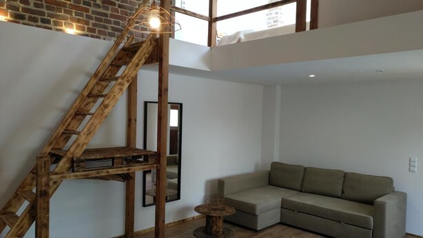 Een loft-achtige woonruimte met een houten trap, een beige bank en een ronde salontafel.