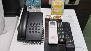 A hotel room with a white bedside table holding a Panasonic phone, a remote control, a menu, and two bottles of water. - Bedbox Hotel (Manila)