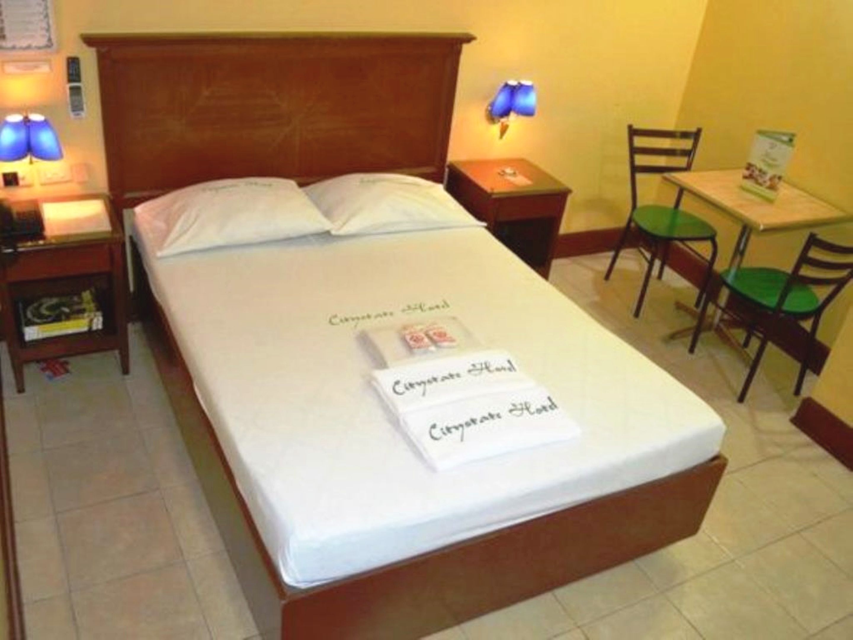 A neatly made bed with a white mattress and two pillows, a wooden headboard, a bedside table with a lamp, a small table with a chair, and a wall-mounted lamp.