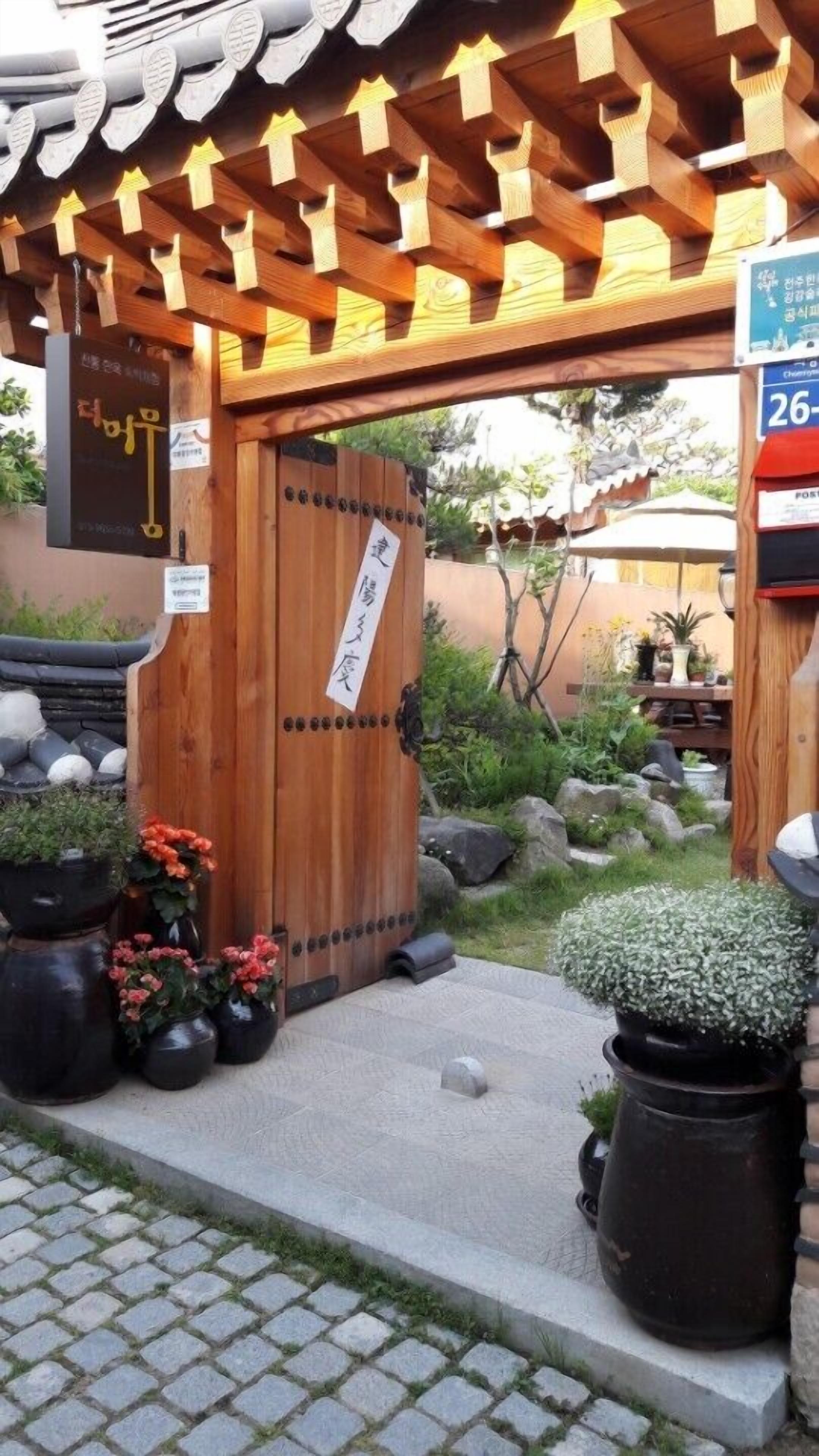 Una puerta de madera tradicional con un letrero, plantas en macetas y un sendero de piedra que lleva a un edificio con techo de tejas.